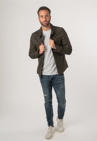 Olivengrüne Wildlederjacke mit Tasche vorne, getragen über einem hellgrauen Hemd, kombiniert mit blauen, verwaschenen Jeans und weißen Sneakers.