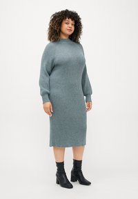Stickad klänning i teal med ribbat mönster, långa ärmar, hög halsringning och rak passform, kombinerad med svarta ankelboots.