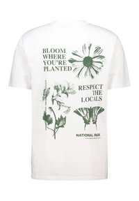 White cotton t-shirt featuring green botanical prints and text: “BLOOM WHERE YOU'RE PLANTED” and “RESPECT THE LOCALS.”