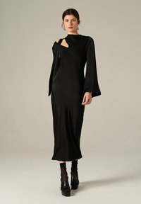 Femme portant une longue robe noire avec des découpes asymétriques et des manches évasées, des chaussettes noires et des chaussures à plateforme, se tenant devant un fond uni.