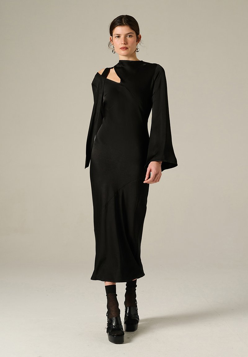 Femme portant une longue robe noire avec des découpes asymétriques et des manches évasées, des chaussettes noires et des chaussures à plateforme, se tenant devant un fond uni.