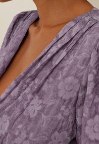 Tissu à motif floral lavande avec une finition douce et texturée. Le vêtement présente un décolleté en V profond et des détails froncés sur les épaules.