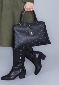 Borsa a tracolla in pelle nera con borchie dorate, forma rettangolare, manici superiori e etichetta staccabile. Indossata con stivali neri fino al ginocchio con bottoni.