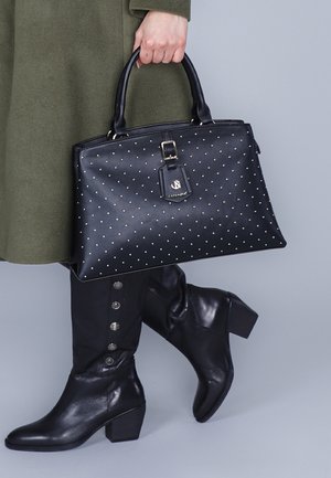 Zwarte leren handtas met gouden studs, rechthoekige vorm, bovenhandvatten en een afneembare tag. Draagt met zwarte kniehoge laarzen met knopen.