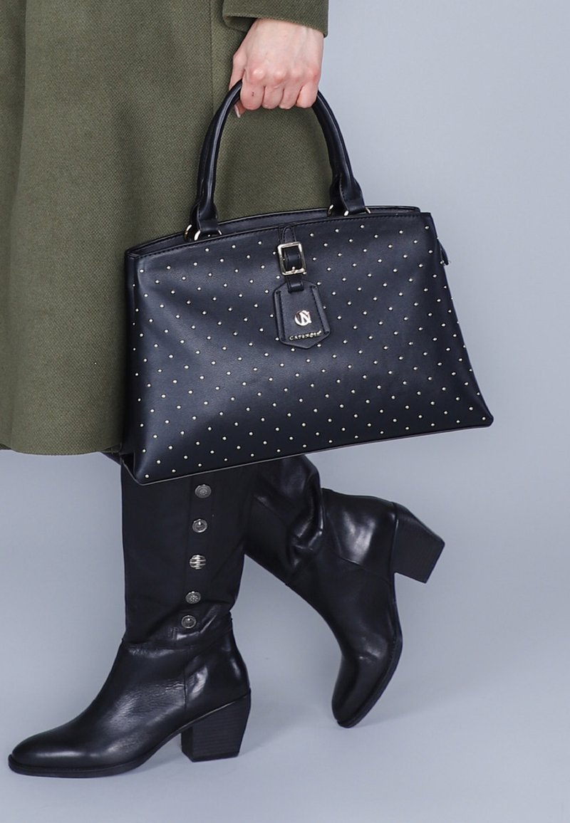 Borsa a tracolla in pelle nera con borchie dorate, forma rettangolare, manici superiori e etichetta staccabile. Indossata con stivali neri fino al ginocchio con bottoni.