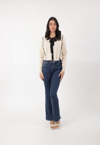 Cárdigan beige con ribete ondulado negro y detalle de lazo, combinado con vaqueros acampanados de mezclilla oscura y sandalias blancas de tiras.