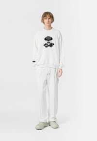 Sudadera blanca con un diseño gráfico negro, combinada con pantalones de chándal blancos. El modelo lleva zapatos de textura clara. Atuendo simple y moderno.