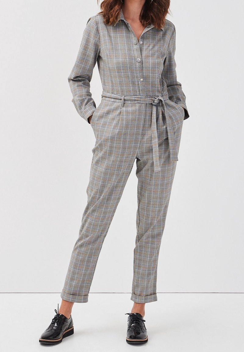 Femme portant une combinaison grise à carreaux avec ceinture, manches longues, et chaussures noires à lacets, debout les mains dans les poches.