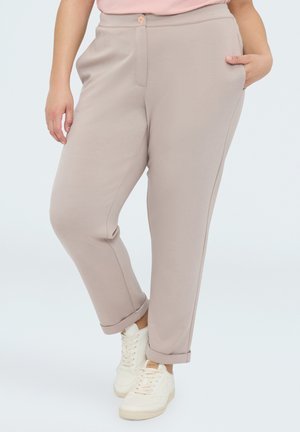 Personne portant un pantalon beige retroussé et des baskets blanches, debout avec une main dans la poche contre un fond clair.