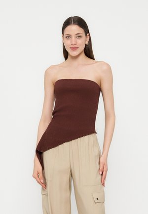 Γυναίκα με ίσια καστανά μαλλιά φοράει ασύμμετρο σκούρο καφέ μπλουζάκι χωρίς τιράντες και φαρδιά μπεζ παντελόνια, στέκεται μπροστά σε απλό λευκό φόντο.