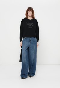 Črna sweatshirt s teksturno grafiko spredaj, v kombinaciji s širokimi modrimi kavbojkami in črnimi špicastimi petami.