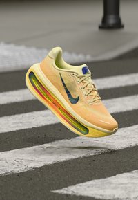 Αθλητικό παπούτσι ανοιχτού κίτρινου χρώματος με επάνω μέρος από διχτυωτό υλικό, πολύχρωμη αερόσολα και σκούρο μπλε λογότυπο σβούρας. Τα κορδόνια και οι ελάχιστες λεπτομέρειες ολοκληρώνουν το σχέδιο.