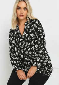 Blusa nera floreale con motivi bianchi, scollatura a V, maniche lunghe e vestibilità comoda. Il tessuto appare leggero e strutturato.