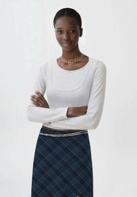 Bluza lungă cu mânecă lungă, de culoare gri deschis, cu decolteu pătrat, asortată cu o fustă cu carouri navy. Fusta are un accent de lanț argintiu la talie.