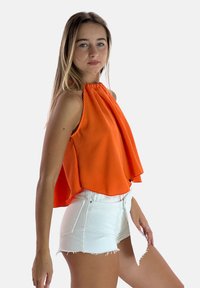 Top arancione brillante senza maniche con un design fluido, abbinato a pantaloni shorts in denim bianco strappati. Abbigliamento estivo semplice e casual.