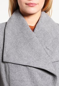 Abrigo de mezcla de lana gris con un gran cuello superpuesto, que presenta una textura suave y líneas limpias. La capa interior visible es de color óxido.