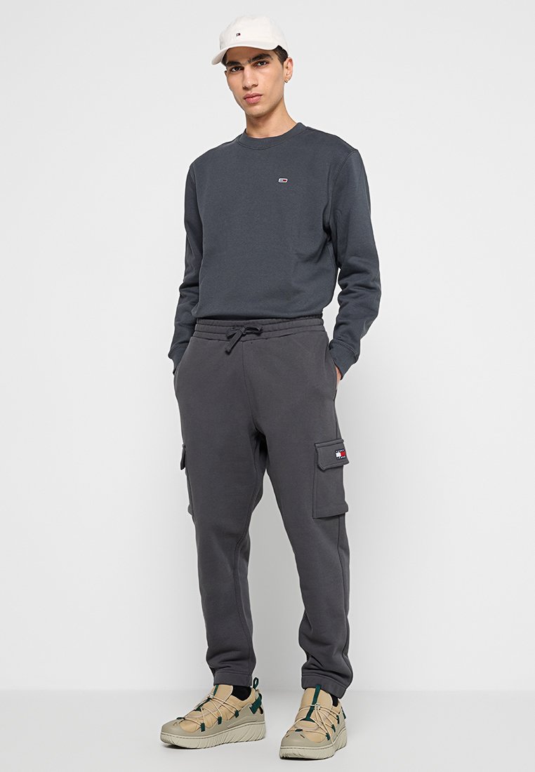 Grå långärmad sweatshirt med liten logga, ihopparad med mörkgrå cargobyxor i sweatmaterial med sidofickor och elastisk midja. Beiga sneakers.