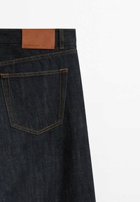 Tummat farkut, joissa on yksi takatasku, kontrastoiva oranssi palautus ompelu ja ruskea nahkaleima vyötäröllä.