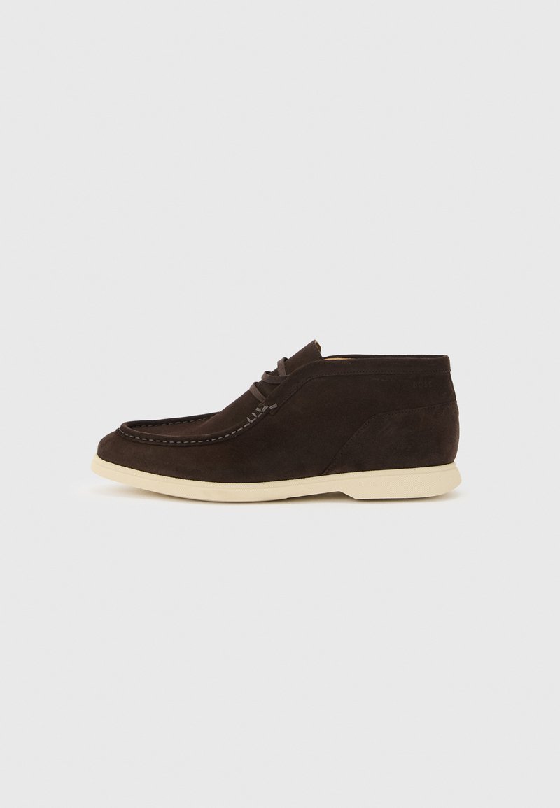 Chaussure décontractée chukka en daim marron foncé avec couture mocassins et semelle en caoutchouc blanche, présentée de profil sur un fond uni.