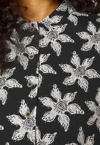 Czarna koszula z dużymi białymi wzorami kwiatowymi z spiralnymi środkami, zapięta przy kołnierzu, z widocznymi kręconymi ciemnymi włosami.