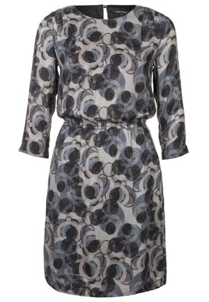 Femme portant une robe mi-longue à manches longues avec des motifs circulaires gris et noirs, des bottines noires et tenant un sac à main noir.