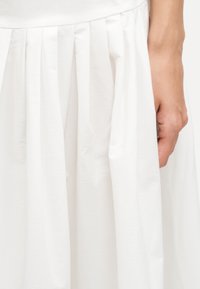 Κοντινό πλάνο μιας λευκής πλισέ φούστας με το χέρι να ακουμπάει δίπλα της, αναδεικνύοντας την υφή του υφάσματος και τις λεπτομέρειες του ενδύματος.