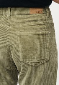 Εγγύς λήψη πίσω τσέπης παντελονιού από ελαφρώς πράσινο κοτλέ ύφασμα με εμφανείς ραφές και καφέ δερμάτινο ταμπελάκι με την επιγραφή "ROXY" στη μέση.