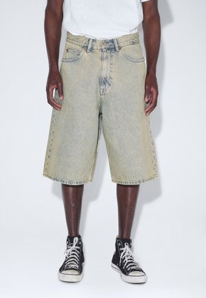 Persoon die lichtgewassen denim knielange shorts draagt, een wit T-shirt en zwarte hoge sneakers met witte veters, staand tegen een effen achtergrond.
