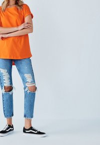 Oranžové bavlněné tričko spárované s roztrhanými světle modrými džíny s roztrhanými okraji. Černé tenisky s bílými akcenty dotvářejí outfit.