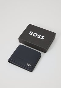Jūras spēku ādas maks ar teksturētu virsmu un sudraba logotipu, novietots blakus melnai kastītei ar uzrakstu "BOSS" treknrakstā baltām burtiem.