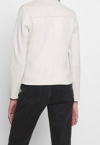 Jasnobeżowa zamszowa kurtka z prostym kołnierzem, długimi rękawami i widocznymi przeszyciami. Noszona z ciemnymi dżinsami, podkreślając smukłą sylwetkę.