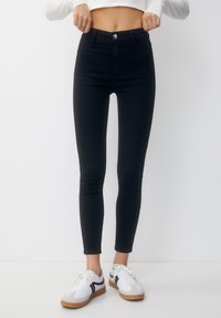 Persona que lleva jeans skinny negros de cintura alta y zapatillas blancas con rayas negras, ajustándose la cinturilla frente a un fondo blanco.
