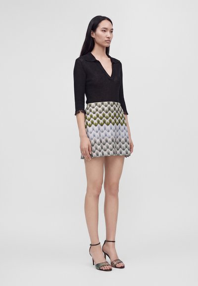 Femme debout portant un haut noir à col en V avec des manches trois-quarts et une jupe courte avec un motif ondulé vert, bleu et gris, ainsi que des sandales à talons hauts.