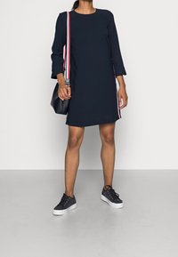 Marineblaues Langarmkleid mit geradem Schnitt, ausgestattet mit rot-weiß gestreiften Seitenakzenten. Kombiniert mit dunklen Sneakern und einer kleinen schwarzen Tasche.