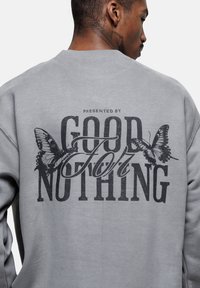 Felpa grigia con grande scritta nera "GOOD FOR NOTHING" e grafiche di farfalle. Realizzata in morbido tessuto, con design a girocollo.