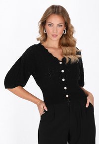 Frau mit welligem blonden Haar, die eine schwarze, knopfverschlossene Strickjacke mit Puffärmeln und schwarze Hosen trägt und mit den Händen in den Taschen posiert.