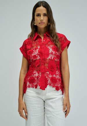 Mujer con una blusa de encaje floral roja de manga corta sobre una camiseta blanca y pantalones blancos, de pie frente a un fondo liso de color gris claro.