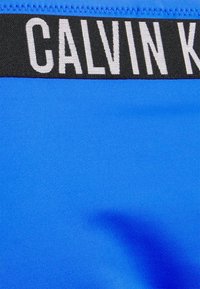 Calções de banho azuis feitos de um tecido suave, com um cós elástico preto onde está impresso "CALVIN KLEIN" em branco.