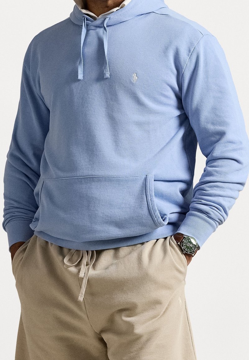 Man wearing light blue hoodie and beige drawstring pants with hands in pockets, also wearing a brown leather-strapped wristwatch with a green face.