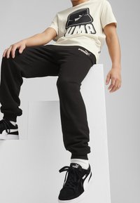 Černé tepláky s bílým logem PUMA, v kombinaci se béžovým tričkem s černou grafikou PUMA. Černé tenisky s bílými akcenty.