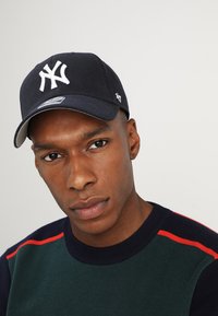 Marineblaues Baseballcap mit weißem gesticktem Logo, gebogenem Schild und grauer Unterseite. Getragen zu einem grünen Pullover mit roten Akzenten.