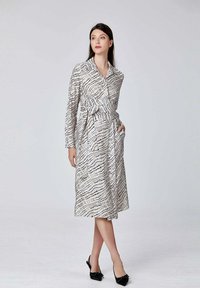 Robe à manches longues, longueur genoux, blanche avec un motif à carreaux noirs, dotée d'une taille ceinturée et de poches latérales. Tissu lisse avec un léger brillant.