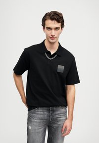 Crna polo majica od pamuka s ovratnikom, s kvadratnom oznakom na lijevoj strani prsiju i kratkim rukavima. Dizajn dodatno naglašava srebrni lančić.