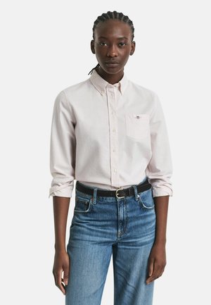 Young woman with braided hair wearing a pale pink button-up shirt with rolled sleeves, tucked into high-waisted blue jeans with a black belt.
