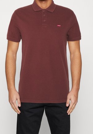 Mand iført en brun kortærmet polo med et lille rødt Levi's-logo på brystet og sorte bukser.