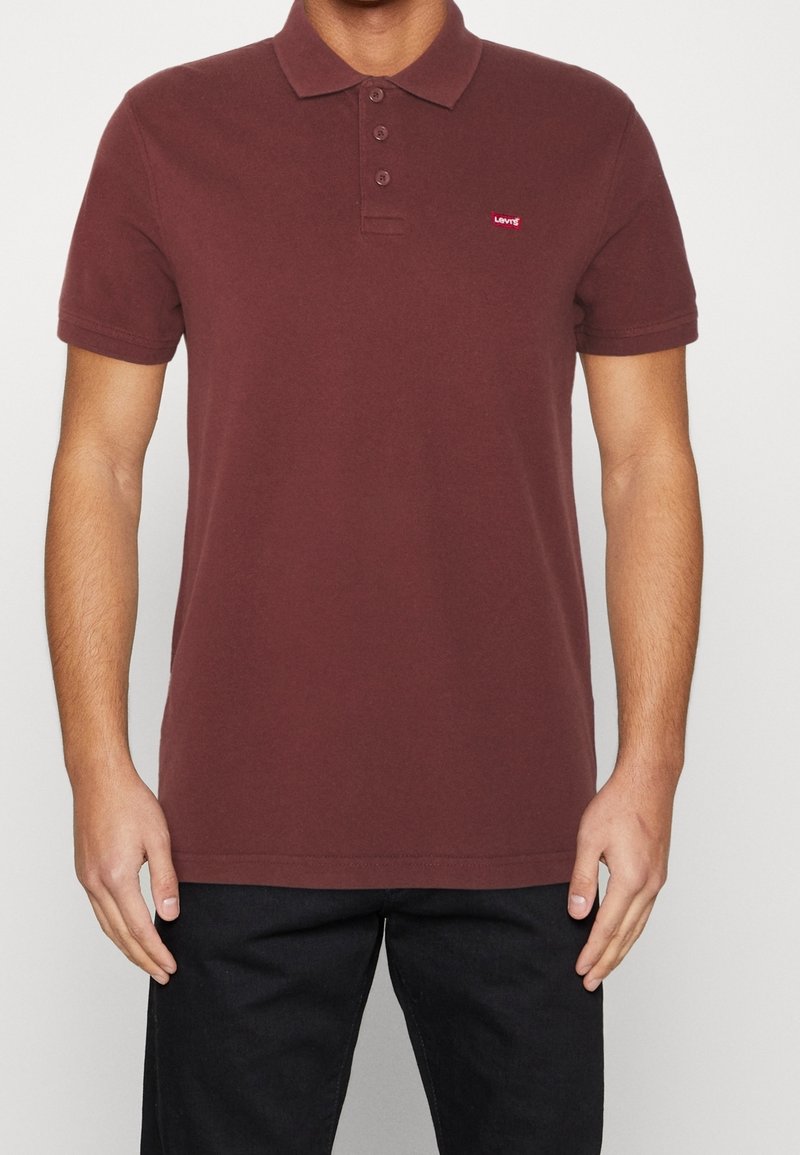 Homme portant un polo marron à manches courtes avec un petit logo rouge Levi's sur la poitrine et un pantalon noir.