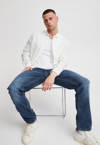 Hvit skjorte over en hvit T-skjorte, kombinert med blå denimjeans og hvite joggesko, sittende på en gjennomsiktig stol.