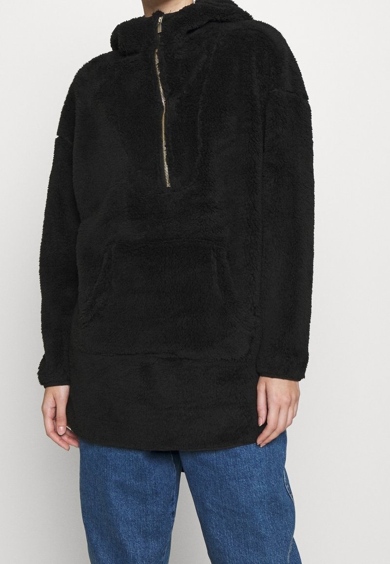 Personne portant un sweat à capuche à demi-fermeture éclair noir en tissu pelucheux avec une grande poche frontale et un jean en denim bleu, sur un fond uni.