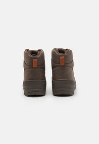 Botas marrones de media altura, hechas de un material texturizado, con un cuello acolchado y un acento de pestaña marrón. Suela de goma con patrones de tracción.