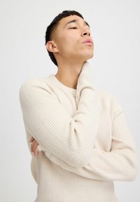 Lichtbeige ribbelgebreide trui met een ronde halslijn en lange mouwen. Het model poseert met een hand op zijn nek.